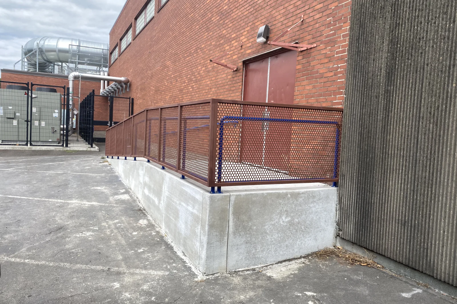 Commercial concrete ADA access ramp featuring bronze expanded metal infill guardrails and blue safety handrails leading to a brick building entrance.