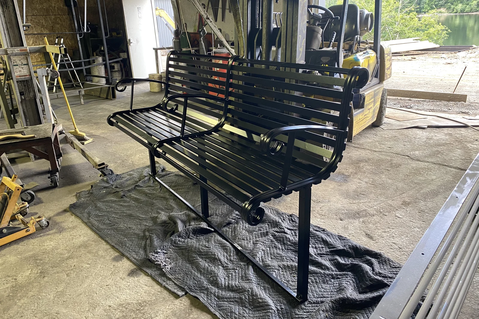 A custom-handcrafted black heavy-duty steel bench with horizontal slats, scrolled armrests, and a central support arm, shown inside the metal fabrication workshop.