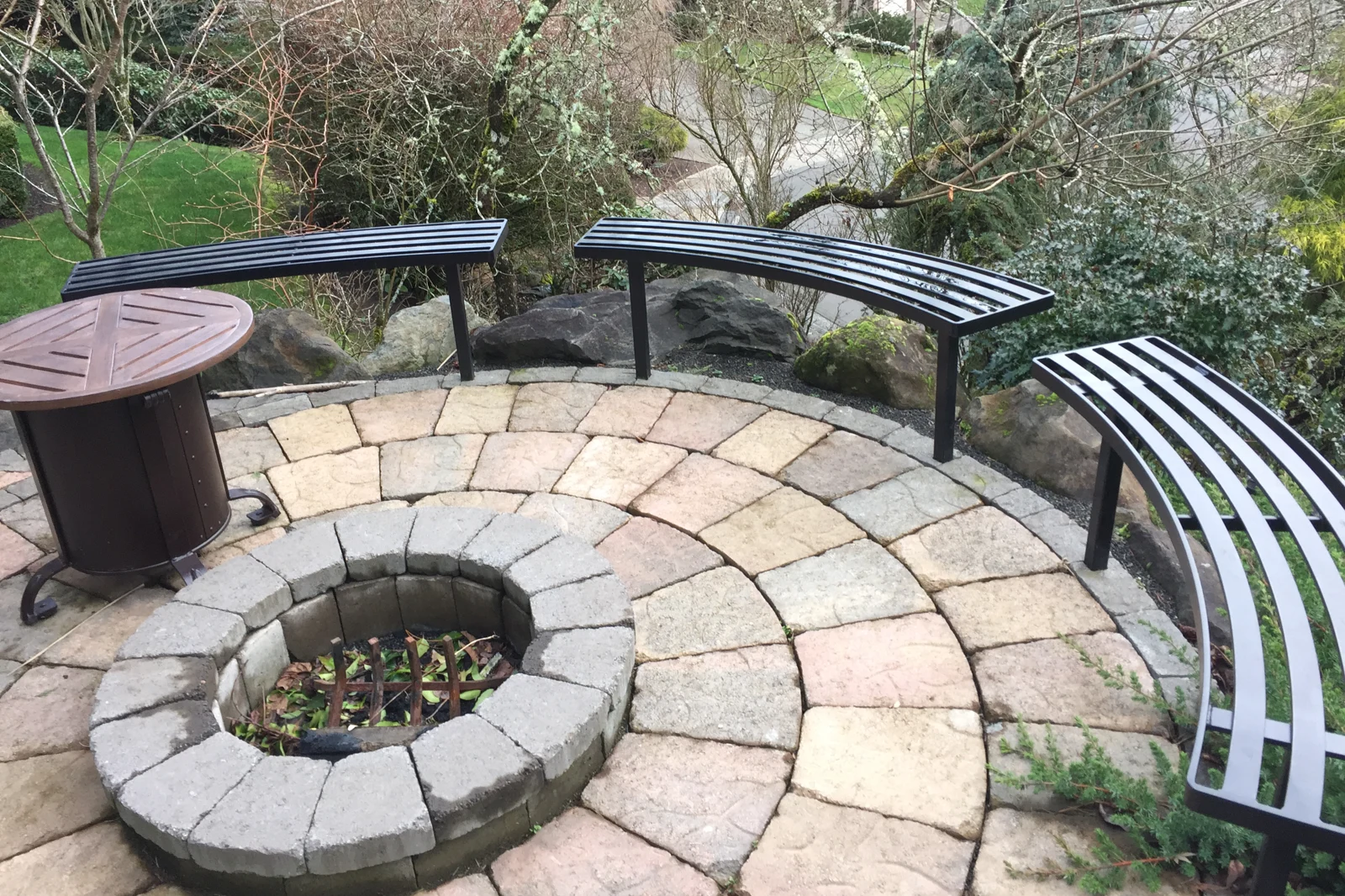 A set of curved black metal benches with a slat design, arranged around a circular stone paver patio and built-in fire pit in a residential backyard.
