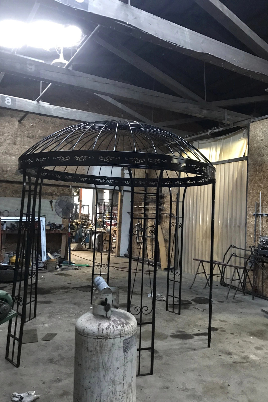 A large, custom-handcrafted black wrought iron gazebo with a dome roof and ornate scrollwork detailing, shown mid-fabrication inside the metalworking workshop.