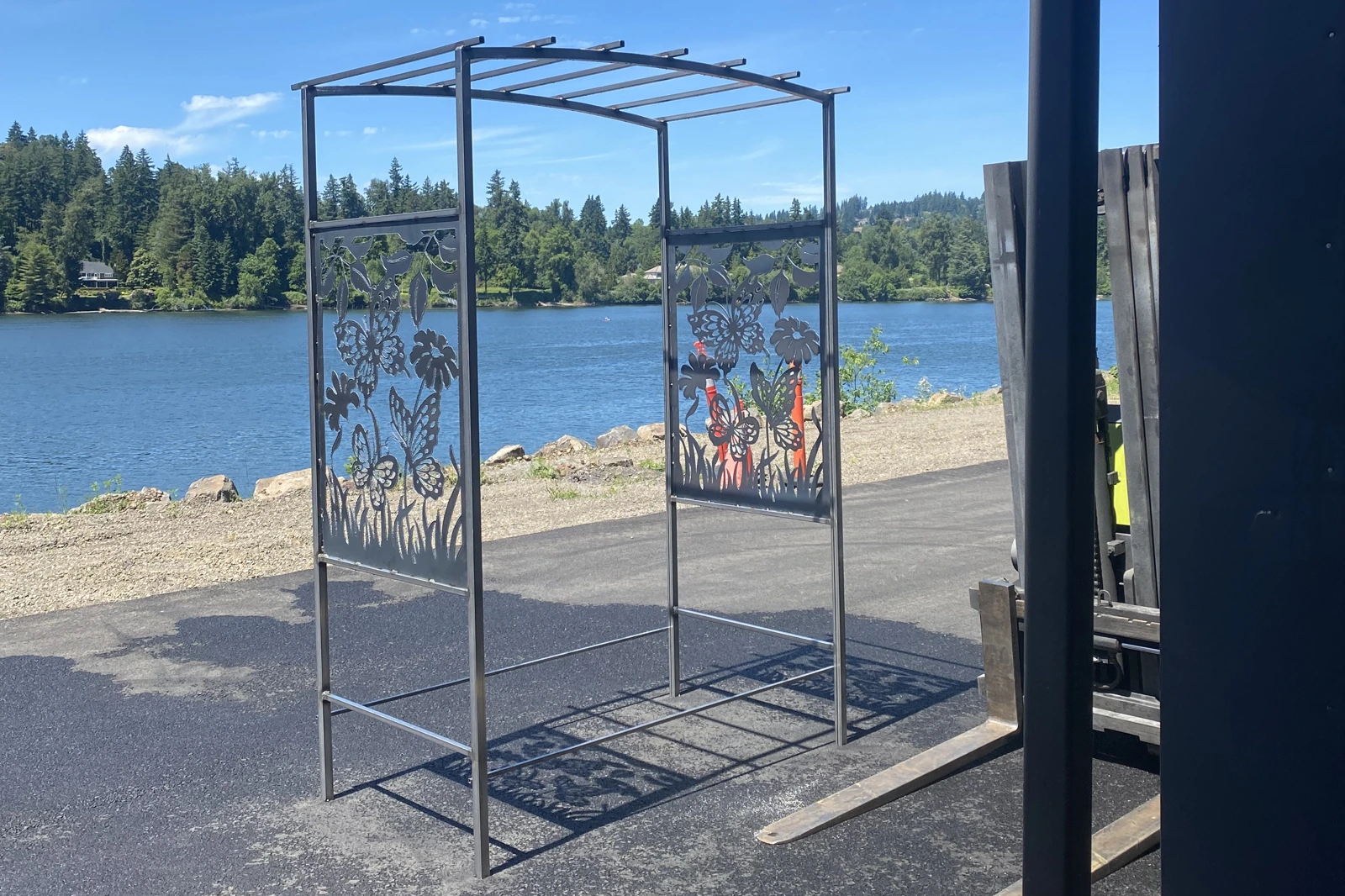A custom-handcrafted black metal garden arbor featuring two decorative side panels with intricate plasma-cut designs of butterflies and wildflowers, set against a scenic lakeside background.