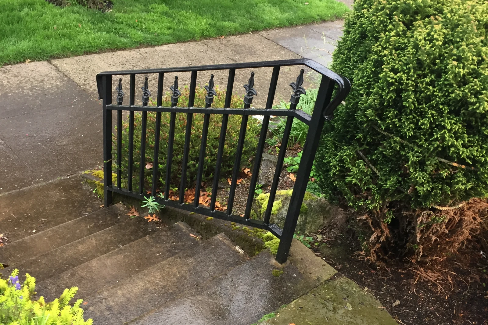 A custom-handcrafted black wrought iron stair railing with decorative fleur-de-lis finials, installed alongside concrete residential steps.