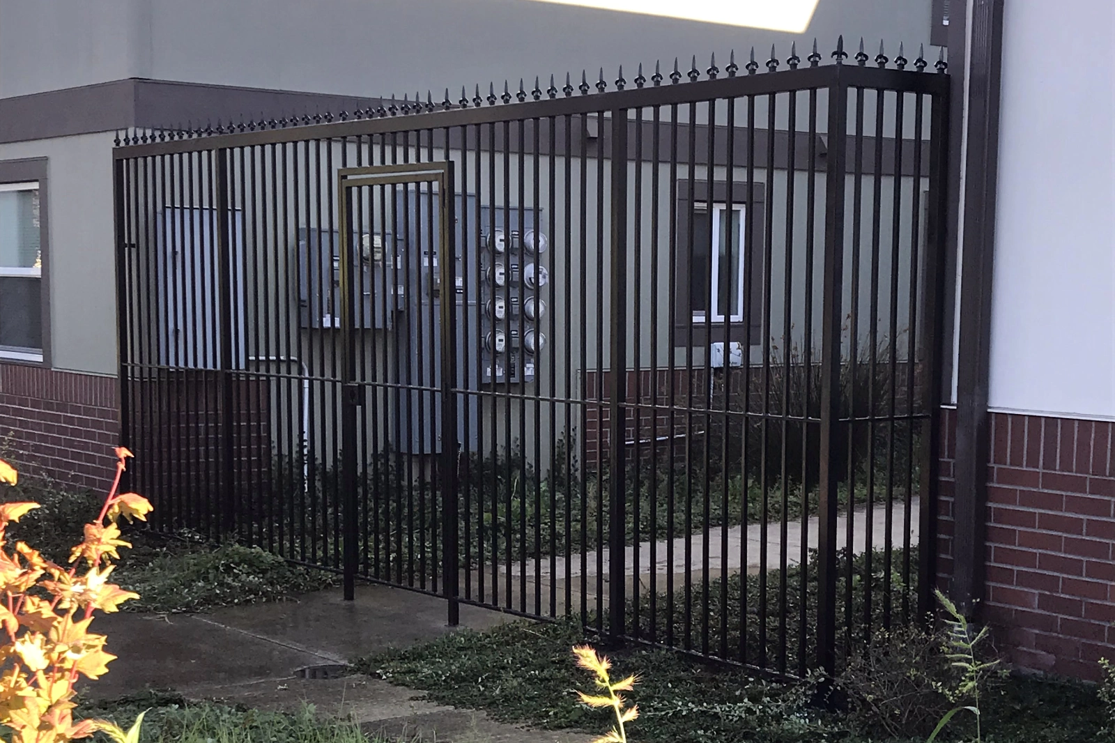 A custom-fabricated tall black steel security fence with pointed finials and a pedestrian gate, enclosing an outdoor utility and meter area for a commercial building.