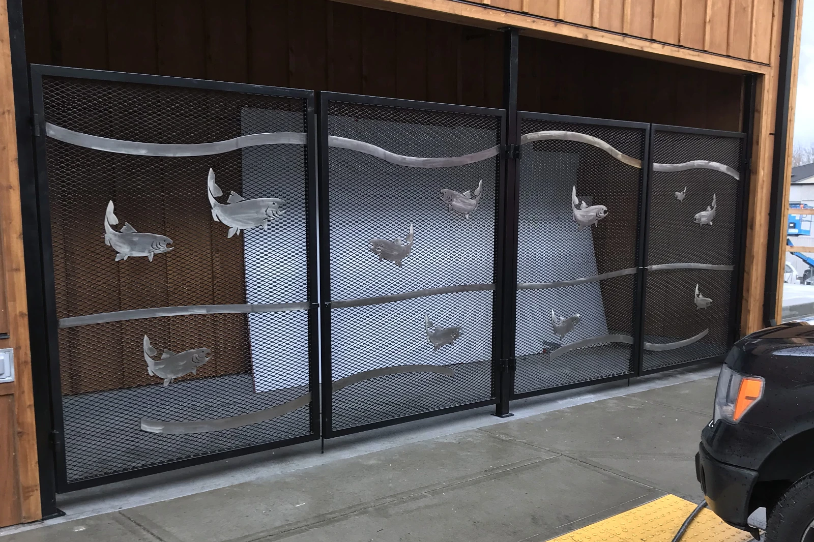 A custom-fabricated black steel mesh security gate featuring artistic brushed metal salmon silhouettes and wavy horizontal accents installed on a commercial building.